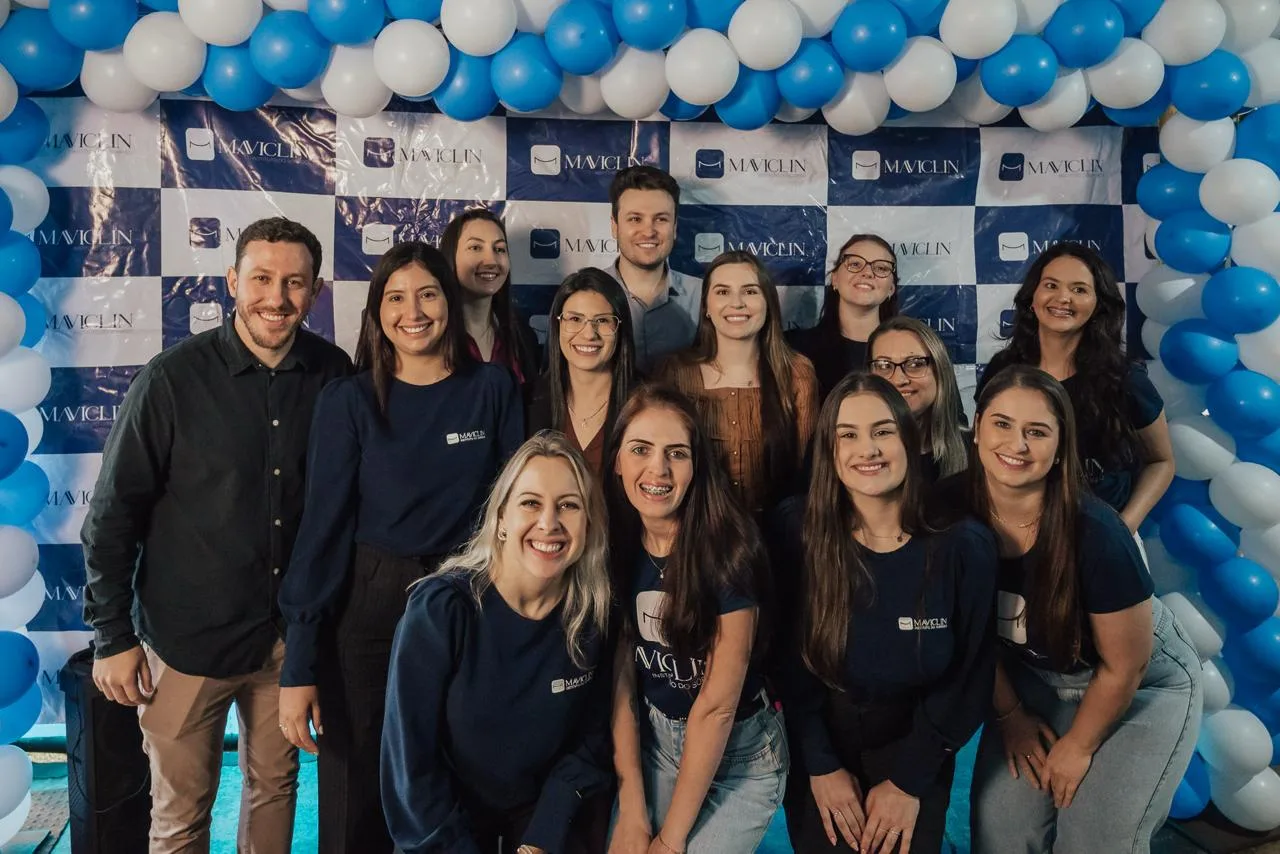 Equipe completa da Maviclin sorrindo, com uniformes da clínica, em frente ao painel com balões azuis e brancos